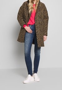Abrigo de estampado de leopardo con un corte holgado, combinado con un suéter rosa brillante y jeans ajustados de color azul marino, completado con zapatillas blancas.
