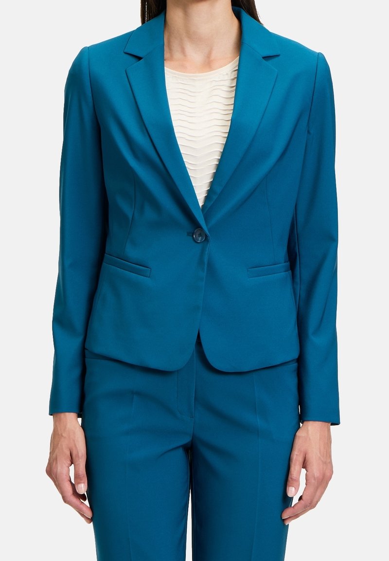 Femme portant un blazer sur mesure couleur turquoise et un pantalon assorti par-dessus un chemisier blanc texturé, debout devant un fond uni.