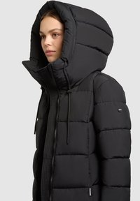 Fekete puffermellény nagy, strukturált kapucnival, steppelt dizájnnal, cipzár elülső résszel és húzózsinóros részletekkel. Simább textúra.