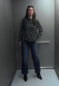 Jeune femme debout dans un ascenseur, portant un chemisier noir à motifs, un jean bleu, des bottines noires et des lunettes de soleil sur la tête.