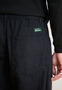 Black cargo pants with an elastic waistband, featuring a back pocket and a green label with mountains. The fabric has a smooth texture.