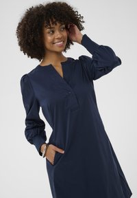 Usmiata žena s kučeravými vlasmi, oblečená v tmavomodrých dlhých šatách, s jednou rukou v vrecku a strieborným náramkom, pohľadom upreným do strany.