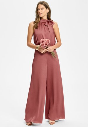 Frau trägt ärmellosen mauvefarbenen Overall mit hohem Kragen und Schleife, hält pinke Blumen, trägt ein goldenes Armband und Creolen, schaut zur Seite.
