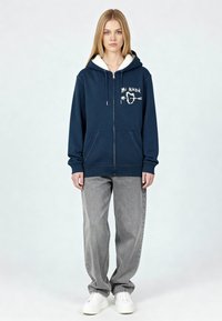 Sweat à capuche zippé bleu marine avec une capuche en polaire blanche, arborant un design brodé "Mi Amor". Assorti avec un jean large gris et des baskets blanches.