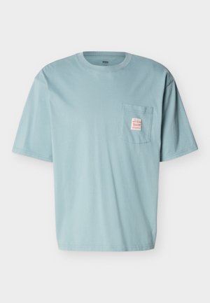 Šviesiai mėlynas trumpomis rankovėmis medvilninis marškinėliai su apvalia apykakle ir krūtinės kišene, ant kurios yra mažas raudonos ir baltos spalvos etikėtė.