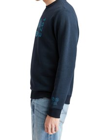 Sudadera azul marino con cuello redondo, mangas largas y un gran diseño gráfico en turquesa en la parte frontal. Puños y dobladillo acanalados.