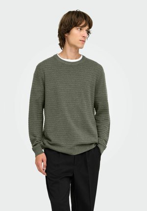 Jeune homme aux cheveux bruns mi-longs portant un pull texturé vert olive et un pantalon noir, regardant vers sa gauche avec les mains dans les poches.