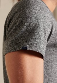 Camiseta gris de manga corta hecha de un tejido texturizado, con una pequeña etiqueta azul de "Superdry" cerca de la costura del hombro.