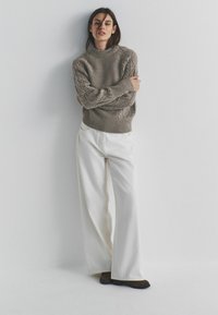 Grijze kabelgebreide trui met een hoge hals, gecombineerd met witte wijde broekspijpen en donkerbruine schoenen, staand tegen een neutrale achtergrond.