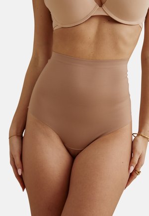 Femme portant un slip gainant taille haute couleur nude sans couture et un soutien-gorge assorti, debout les mains détendues le long du corps.