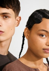 Pendientes de aro de oro llevados por una persona con cabello corto y otra con cabello trenzado. Proximidad cercana, destacando rasgos faciales y accesorios.