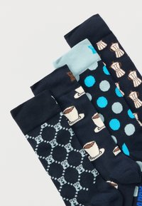 Τέσσερα ζεύγη ναυτικών κάλτσες με σχέδια: κουκουβάγιες, κούπες καφέ, μπλε και γκρι πουά, και μοκάτες, τοποθετημένα επικαλυπτόμενα σε λευκή επιφάνεια.