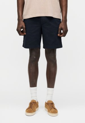 Muž oblečený v béžovej košeli, tmavomodrých šortkách, bielych ponožkách a hnedých teniskách, stojí pred jednoliatym svetlým pozadím.