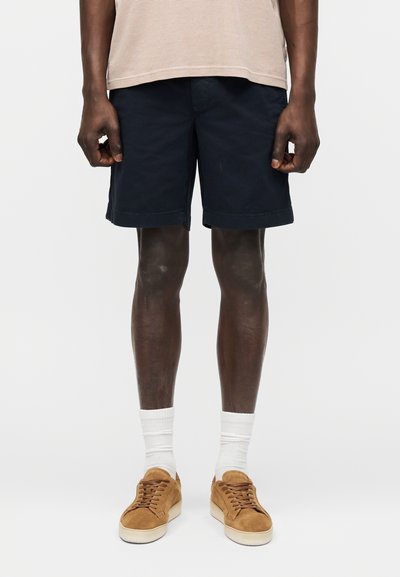 Homme portant une chemise beige, un short bleu marine, des chaussettes blanches et des baskets marron, debout sur un fond clair uni.