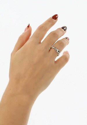 Silberner Metallring mit wellenförmigem Design, getragen an einer Hand mit mehrfarbigen Nägeln. Glatte Textur vor einem schlichten weißen Hintergrund.