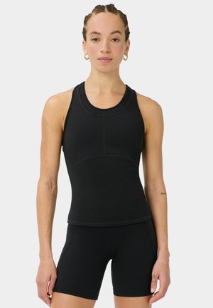 Femme aux cheveux tressés portant un haut de sport noir sans manches et un short assorti, debout devant un fond clair uni.