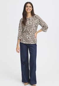 Mosolygó nő hosszú, sötét hajú, mintás barna és fehér toppot, széles lábú sötétkék farmert és bézs hegyes orrú cipőt visel.