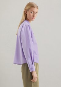 Blusa lilla chiaro con una vestibilità morbida, maniche lunghe, polsini con bottoni e scollatura rotonda, abbinata a pantaloni verde oliva.