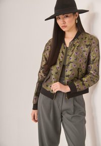 Mujer con cabello largo y oscuro, usando un sombrero negro de ala ancha, una chaqueta bomber floral en verde y morado, pantalones gris de cintura alta y aretes dorados en forma de aro.