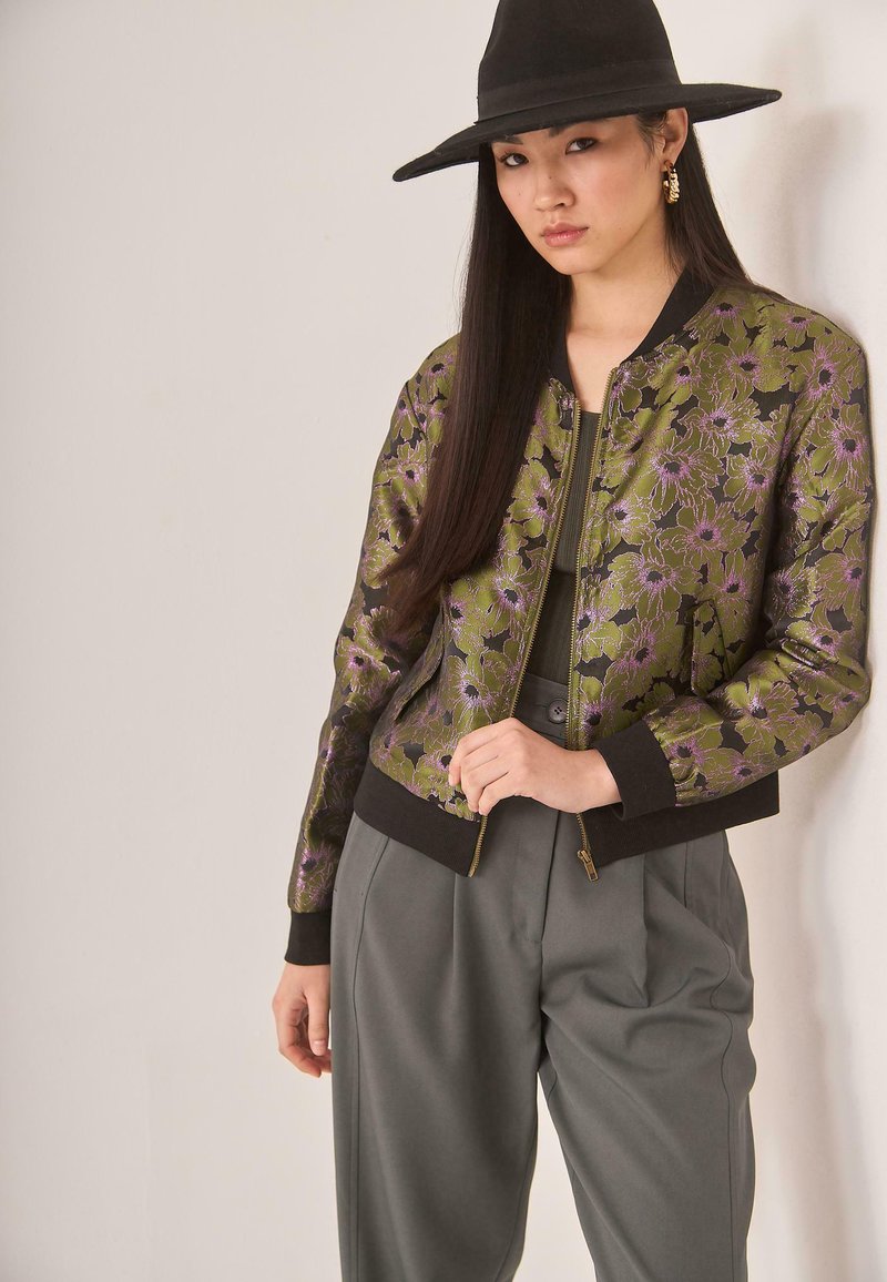Mujer con cabello largo y oscuro, usando un sombrero negro de ala ancha, una chaqueta bomber floral en verde y morado, pantalones gris de cintura alta y aretes dorados en forma de aro.