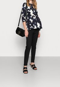 Mujer embarazada vestida con una blusa de patrón negro y blanco, pantalones negros, sandalias de tacón negro y llevando un bolso de hombro negro.