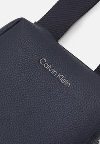Borsa a tracolla in pelle blu navy con superficie testurizzata; presenta il logo grigio "Calvin Klein"; cinturino in tessuto regolabile.