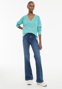 Turquoise knitted sweater with a V-neck and long sleeves, paired with blue flared jeans. Model wears white sneakers, creating a casual look.