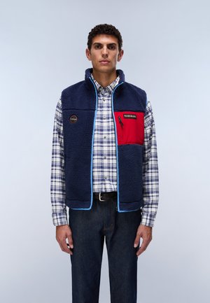 Ein Mann trägt eine marineblaue Fleeceweste mit einer roten Brusttasche über einem weiß-blau karierten Hemd und dunklen Jeans und steht vor einem einfarbigen Hintergrund.