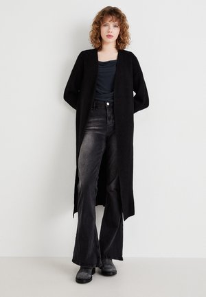 Cardigan lungo nero con apertura frontale e spacco laterale, indossato sopra un top scuro e jeans neri a palazzo. Tessuto morbido e vestibilità comoda.