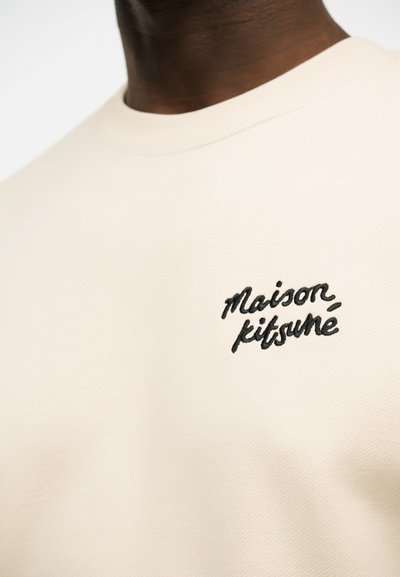 Κοντινό πλάνο ενός ατόμου που φοράει μπεζ φούτερ με στρογγυλή λαιμόκοψη, με την επιγραφή «maison kitsuné» κεντημένη με μαύρα γράμματα στο αριστερό μέρος του στήθους.