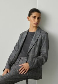 Žena s hladce sčesanými vlasy má na sobě šedý kostkovaný blazer a kalhoty, sedí proti světle šedé zdi, ruce má položené na klíně.