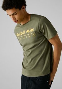 Olivno zelena bombažna majica s kratkimi rokavi, ki na sprednji strani vsebuje rumen logotip Red Bull Racing. Ima gladko teksturo in okroglo ovratnico.