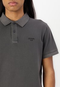 Personne portant un polo gris foncé avec un col, deux boutons et le logo "JOOP! JEANS" sur la poitrine gauche.