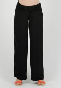 Schwarze, weit geschnittene Hose mit strukturiertem Finish, ausgestattet mit einem gerafften Bund und seitlichen Taschen. Kombiniert mit orangefarbenen Sandalen für einen Kontrast.