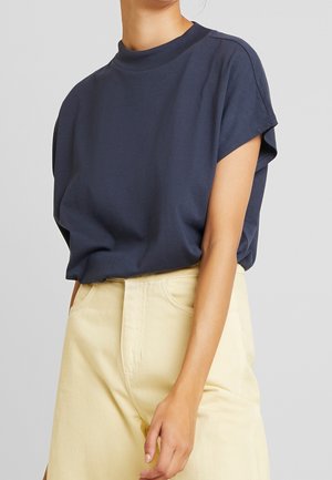 Mørkeblå t-shirt med korte ærmer og høj hals, lavet af blødt stof. Kombineret med lysegule, højtaljede bukser.