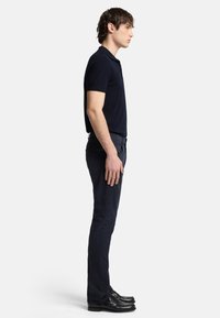 Polo shirt navy a maniche corte abbinato a pantaloni scuri. Il modello è in piedi di lato, mostrando una vestibilità slim e una texture liscia. Scarpe nere completano il look.