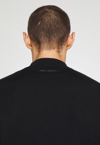 Vue arrière d'une personne aux cheveux courts portant une veste noire côtelée avec un discret logo "KARL LAGERFELD" au niveau de l'encolure.