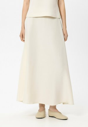 Frau trägt einen langen, cremefarbenen Rock mit passenden cremefarbenen Slip-On-Schuhen und steht vor einem schlichten weißen Hintergrund.