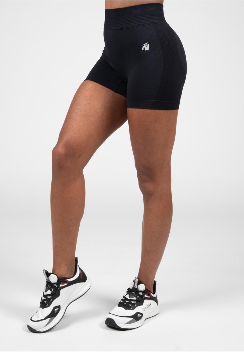 Zwarte sportshorts van rekbaar materiaal met een naadloze tailleband en een klein logo aan de zijkant, gecombineerd met zwart-witte sneakers.