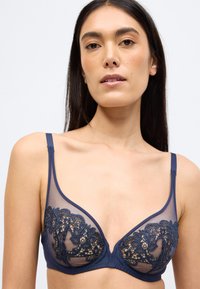 Σουτιέν με μπλε ναυτικό δαντέλα και διαφανείς φάσες από δίχτυ, διακοσμημένο με κεντήματα λουλουδιών και ρυθμιζόμενους ιμάντες. Η απαλότητα της υφής και το τριγωνικό σχήμα αναδεικνύουν το σχέδιο.