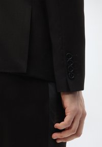 Zwarte op maat gemaakte colbert van een gladde stof, voorzien van een revers, zes knopen op de manchetten en een slanke pasvorm. Een hand bevindt zich dicht bij het colbert.