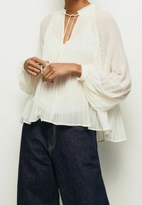 Vit veckad blus med hög krage, knytdetaljer och långa, fluffiga ärmar; bärs tillsammans med mörkblå vida jeans.