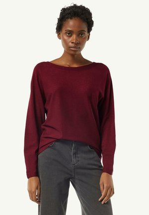 Burgunderfarbener Pullover mit weitem Ausschnitt und langen Ärmeln, aus weichem, strukturiertem Stoff. Kombiniert mit grauen Jeans.