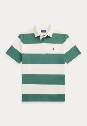 Młody mężczyzna w zielono-białej paskowanej koszulce polo, białych szortach, białych tenisówkach, zielonej czapce i ciemnym swetrze zarzuconym na ramiona.