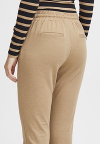 Bruna sweatpants i en texturerad tyg, elastisk midja och två bakfickor. Bilden visar byxorna bakifrån med en modells hand vilande i närheten.