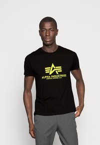 Black short-sleeve t-shirt featuring a large yellow "Alpha Industries" logo with geometric accents. Smooth fabric texture.