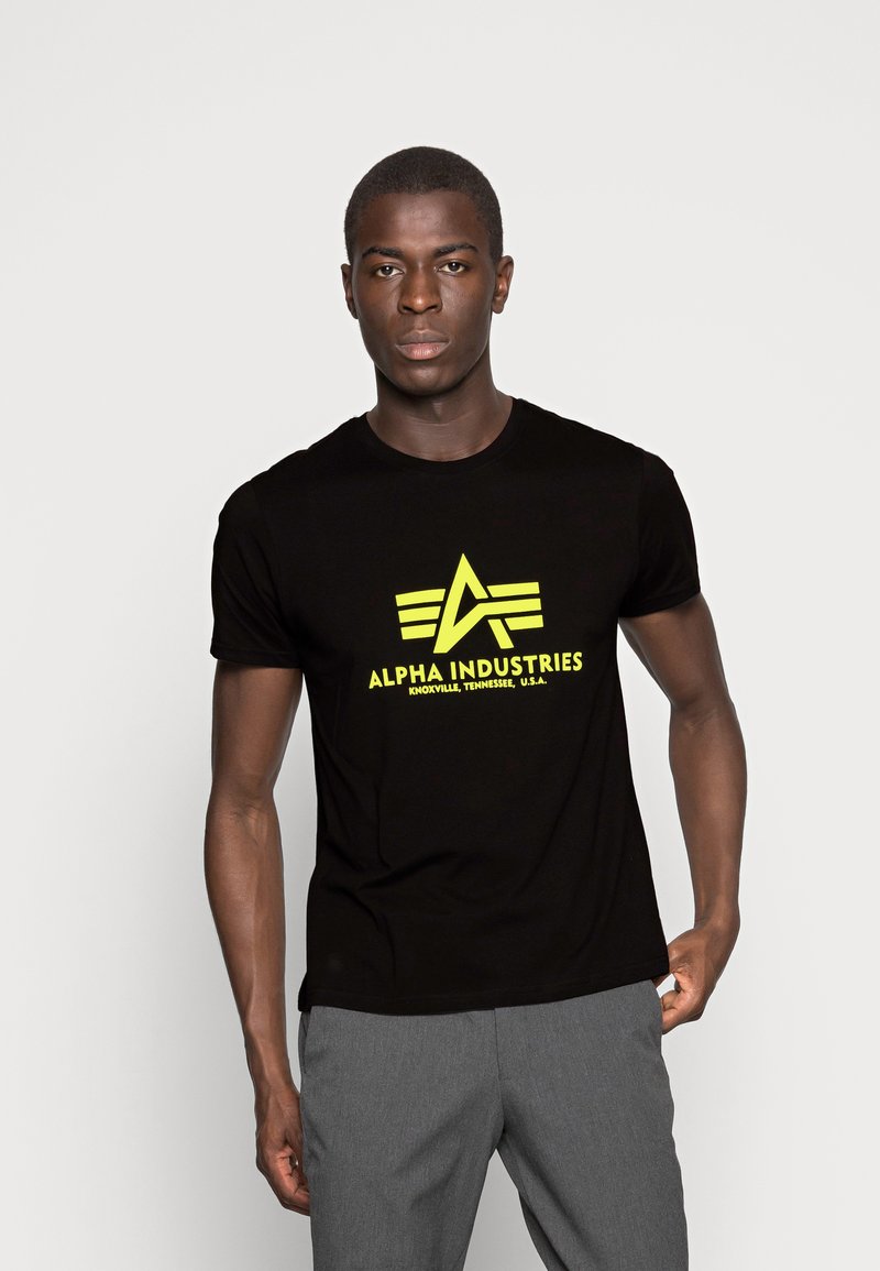 Black short-sleeve t-shirt featuring a large yellow "Alpha Industries" logo with geometric accents. Smooth fabric texture.