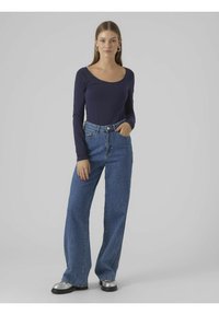 Marineblaues Langarmoberteil mit weitem Halsausschnitt, kombiniert mit hochgeschnittenen, locker sitzenden blauen Jeans. Das Model trägt silberne Plateauschuhe.