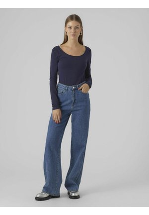 Marineblå langærmet top med en bred halsudskæring, parret med højtaljede, løse blå jeans. Modellen bærer sølvfarvede plateau-sko.