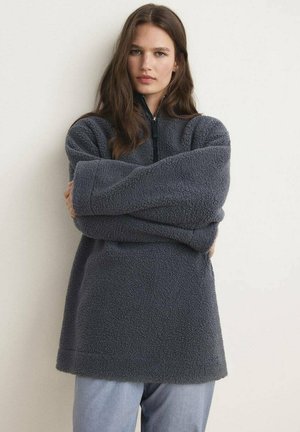 Pullover grigio in fleece con colletto alto e zip a metà. Presenta maniche oversize e una finitura morbida e testurizzata.